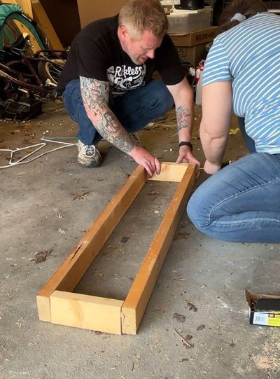 Tom holding framing 2x4 pieces together on a garage floor while Em places a screw